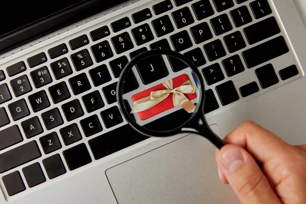 Vista recortada del hombre sosteniendo lupa por encima del teclado del ordenador portátil con regalo, concepto de compras de vacaciones en línea - foto de stock