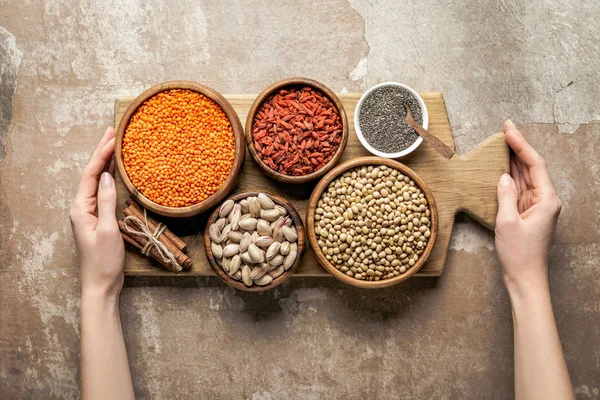 Vista cortada de mulher segurando placa de madeira com legumes e goji bagas no fundo rústico — Fotografia de Stock