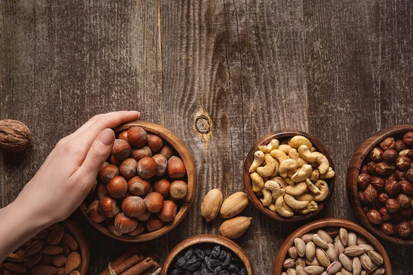 Vista parcial de la mujer sosteniendo tazón con avellanas en la mesa de madera con diferentes nueces alrededor - foto de stock