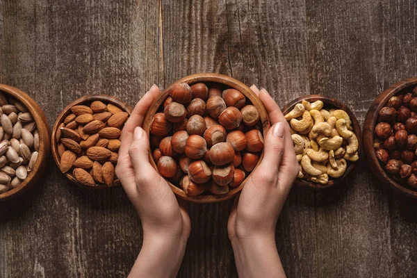 Vista parziale della donna che tiene ciotola con nocciole su un tavolo di legno con diverse noci intorno — Foto stock