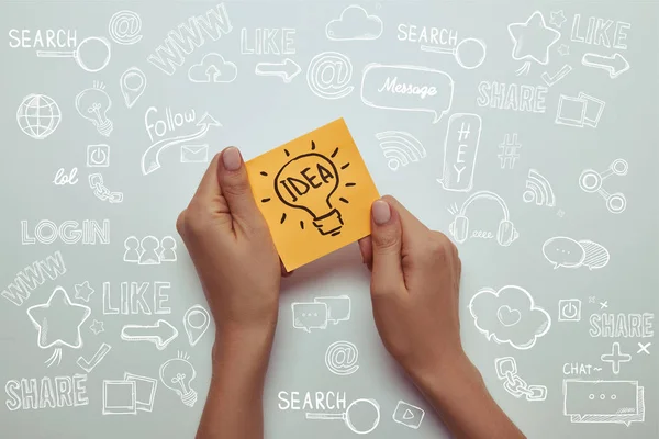 Cropped image of woman holding paper sticker with word idea in lightbulb on white with marketing icons — Stock Photo