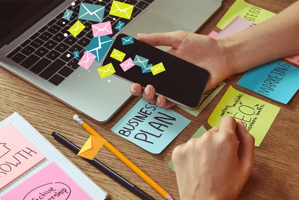 Cropped image of woman holding smartphone with email icons at table with business plan sticker — Stock Photo