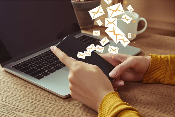 Imagen recortada de la mujer sosteniendo teléfono inteligente con iconos de correo electrónico en la mesa con el ordenador portátil - foto de stock