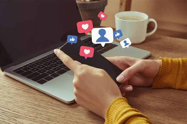 Cropped image of woman holding smartphone with multimedia icons at table with laptop — Stock Photo