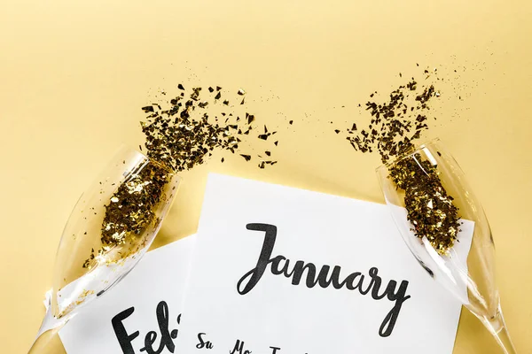 Copos de champanhe com confete dourado, papéis com calendário de janeiro e fevereiro em fundo bege — Fotografia de Stock