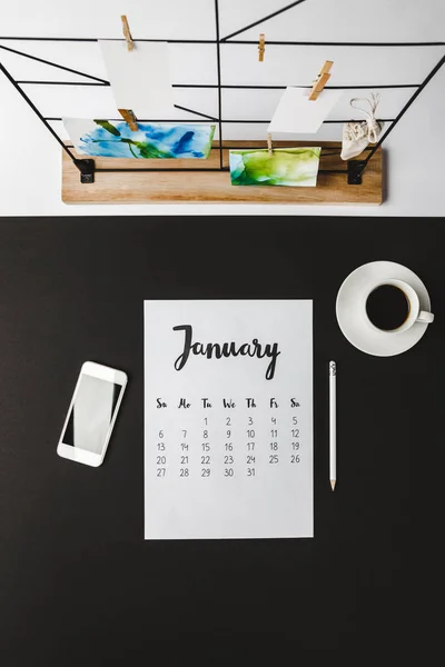 Vue du haut du calendrier de janvier, crayon, smartphone avec écran vierge et tasse de café sur noir — Photo de stock