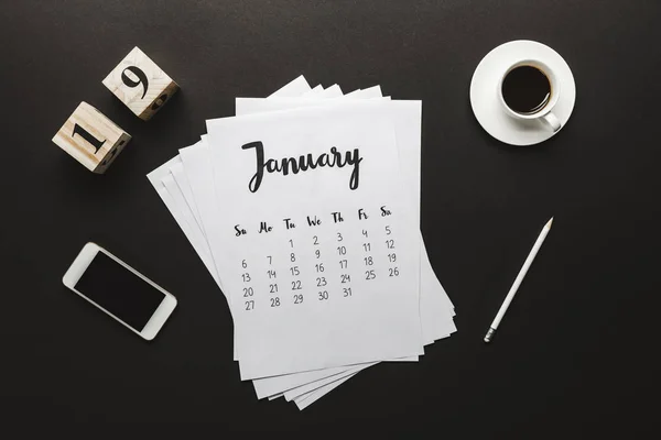 Top view of calendar, wooden cubes with 19 numbers, pencil, cup of coffee and smartphone on black — Stock Photo