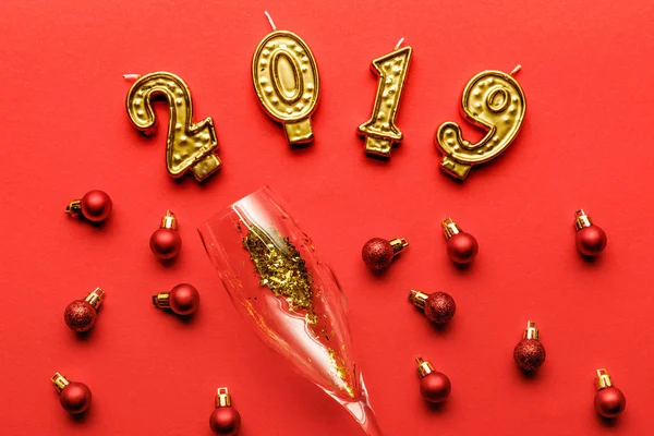 Top view of champagne glass with golden confetti, 2019 candles and red christmas baubles on red — Stock Photo