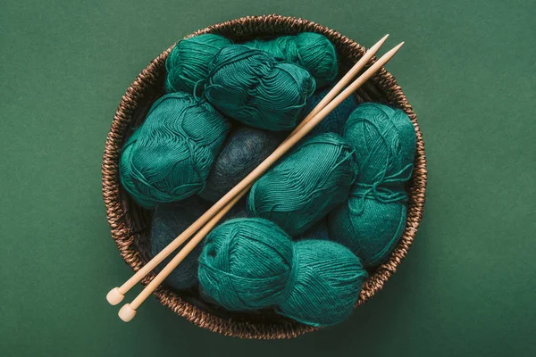 Top view of green and blue yarn clews and knitting needles in wicker basket on green backdrop — Stock Photo