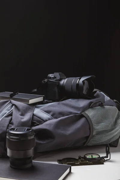 Sac à dos, appareil photo avec objectif, cahiers avec stylo et boussole sur noir, concept de voyage — Photo de stock