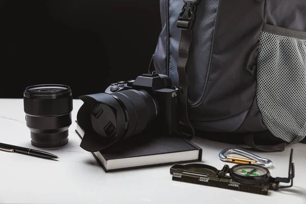 Vista de cerca de la cámara fotográfica con lente, bolígrafo, cuaderno, brújula y mochila en negro - foto de stock