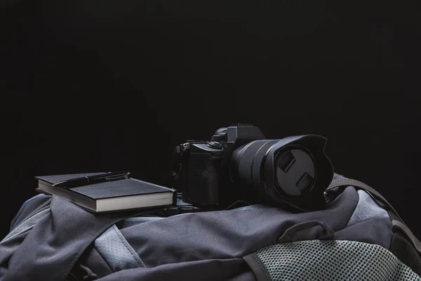 Зменшеному вигляді фотоапарат з записну книжку й ручку на рюкзак ізольовані на чорному — стокове фото