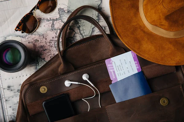 Top view of leather bag with passport, ticket, smartphone with earphones, sunglasses, lens and hat on map — Stock Photo