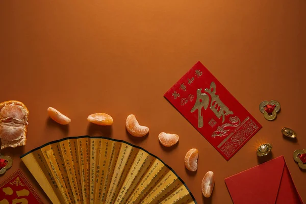 Top view of peeled tangerine, fan with hieroglyphs and red envelope on brown background — Stock Photo