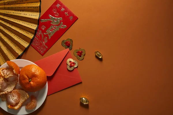 Mandarinen auf Teller, Ventilator mit Hieroglyphen, goldenen Dekorationen und rotem Umschlag auf braunem Hintergrund, chinesische Neujahrskomposition — Stockfoto