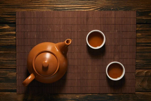 Vista superior de la tetera de cerámica marrón y tazas de té en la estera de bambú - foto de stock