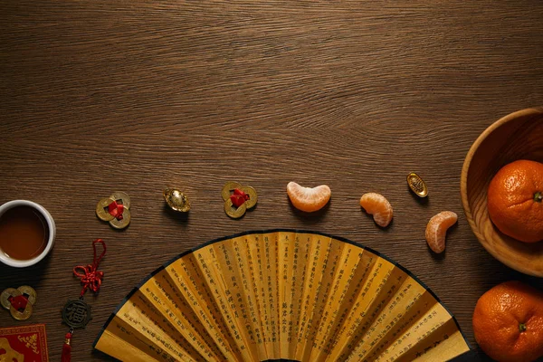 Vue de dessus de mandarines fraîches mûres, tasse de thé, ventilateur avec hiéroglyphes et pièces d'or sur table en bois — Photo de stock