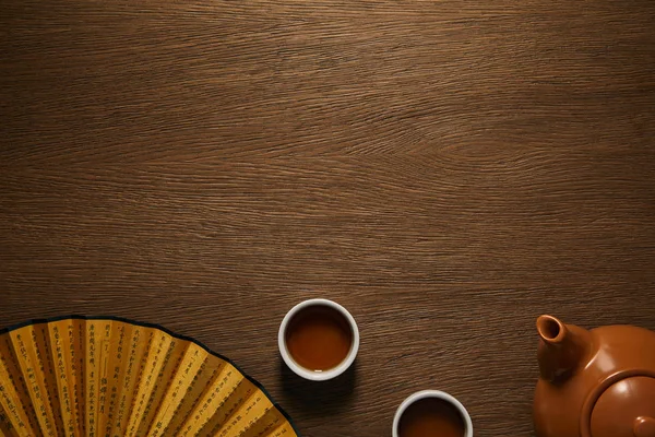 Top view of golden fan with hieroglyphs and teapot with cups on wooden background — Stock Photo