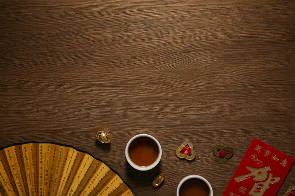 Vue du dessus des tasses à thé, ventilateur avec hiéroglyphes et pièces d'or sur la surface en bois — Photo de stock