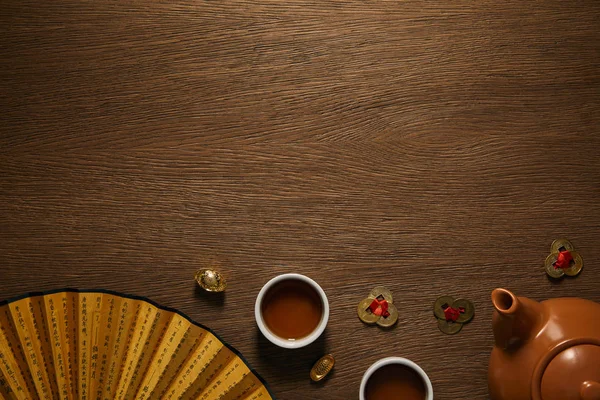 Top view of tea set, fan with hieroglyphs and golden coins on wooden surface — Stock Photo