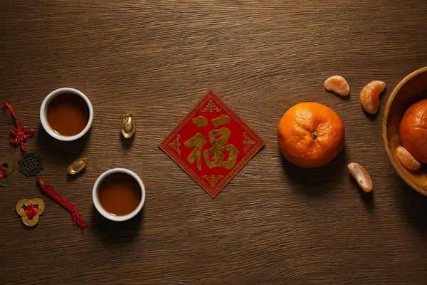 Top view of fresh ripe tangerines, golden hieroglyph, cups of tea and coins on wooden surface — Stock Photo