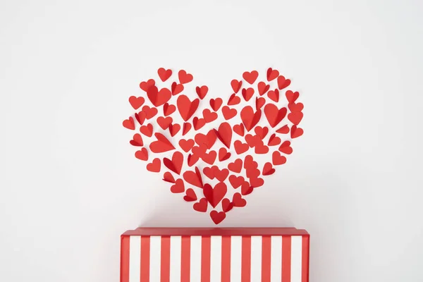 Vista superior de la disposición en forma de corazón de pequeños corazones cortados de papel rojo y caja de regalo a rayas sobre fondo blanco - foto de stock