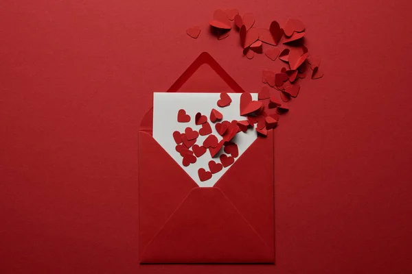 Top view of white letter in envelope with pile of paper cut hearts on red background — Stock Photo