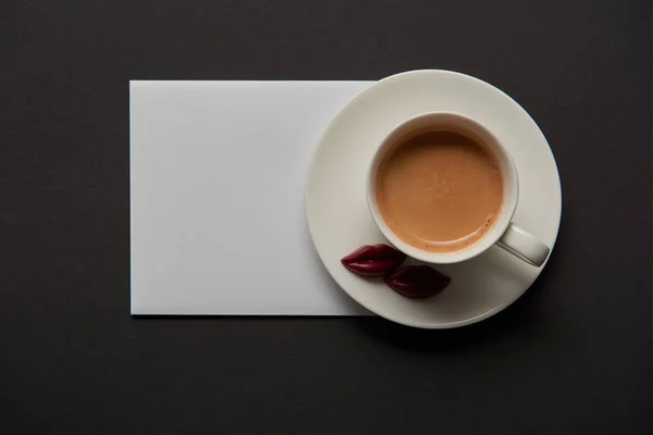 Top view of cup with coffee, chocolate lips on saucer and empty white card on black background — Stock Photo