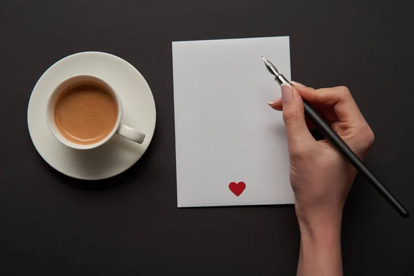 Vista recortada de la mujer escribiendo en la tarjeta de felicitación vacía con signo de corazón rojo cerca de la taza de café - foto de stock