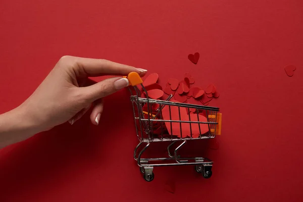 Vista recortada de la mano femenina y el carrito de compras decorativo con corazones cortados en papel sobre fondo rojo - foto de stock