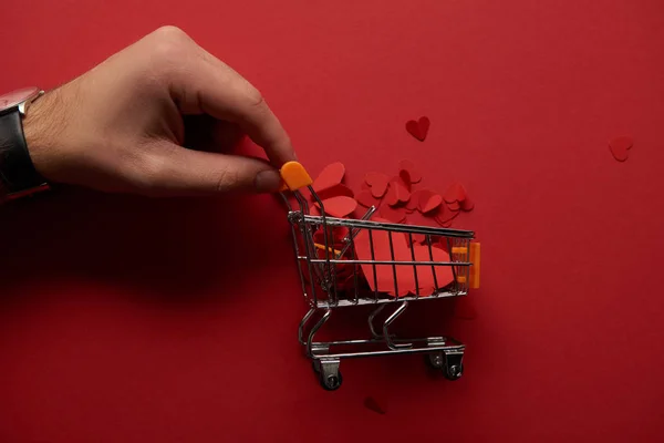 Vista recortada de mano masculina y carrito de compras de juguete con corazones cortados en papel sobre fondo rojo - foto de stock