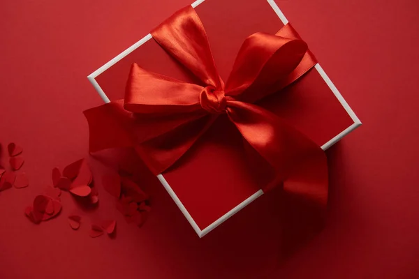 Верхний вид красного подарка с шелковой лентой и разрезанными бумагой сердцами на красном фоне — стоковое фото