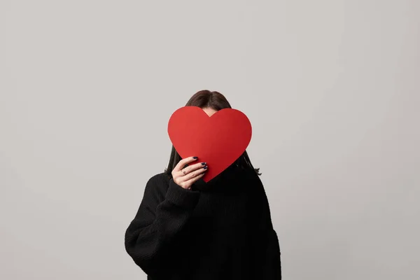 Mujer joven en negro con cara oscura y papel vacío tarjeta de corazón cortado aislado en gris - foto de stock