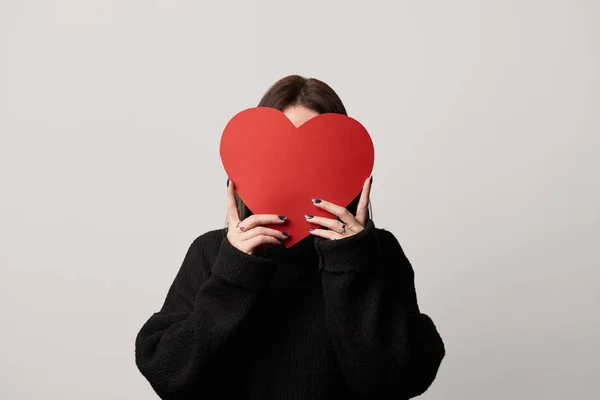 Chica en negro con cara oscura y papel vacío tarjeta de corazón cortado aislado en gris - foto de stock