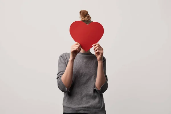 Mujer ocultar la cara detrás de papel en blanco cortar la tarjeta del corazón aislado en gris - foto de stock