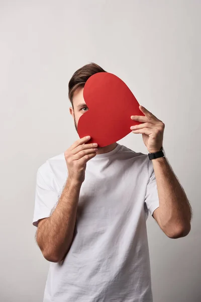 Hombre ocultar la cara detrás de papel en blanco tarjeta de corazón cortado sobre fondo gris - foto de stock