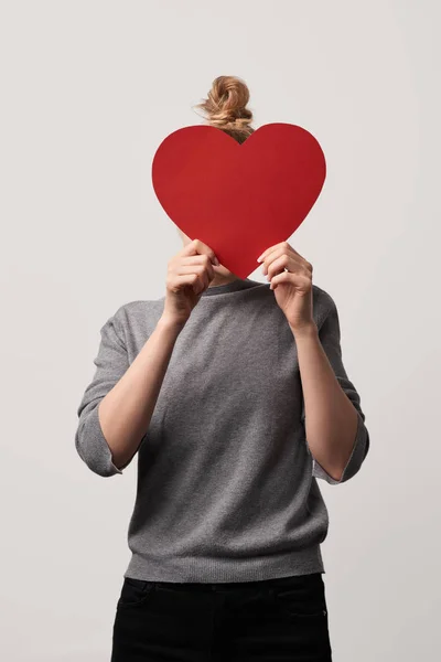 Chica ocultar la cara detrás de papel en blanco cortar tarjeta de corazón aislado en gris - foto de stock