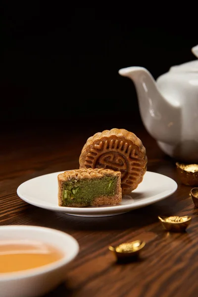 Selective focus of traditional chinese mooncakes, tea pot and gold ingots on wooden table isolated on black — Stock Photo