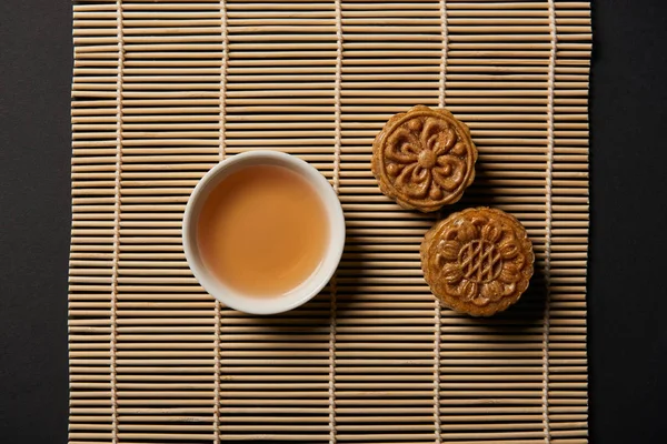 Vista superior de tortas de luna chinas y taza de té en la estera de mesa de bambú - foto de stock