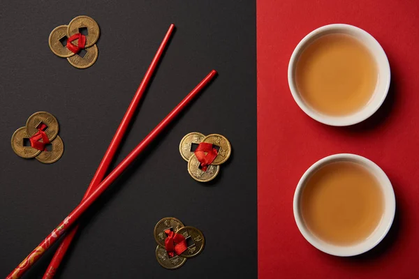 Vue du haut des baguettes avec du thé chinois traditionnel et des pièces de feng shui sur fond rouge et noir — Photo de stock