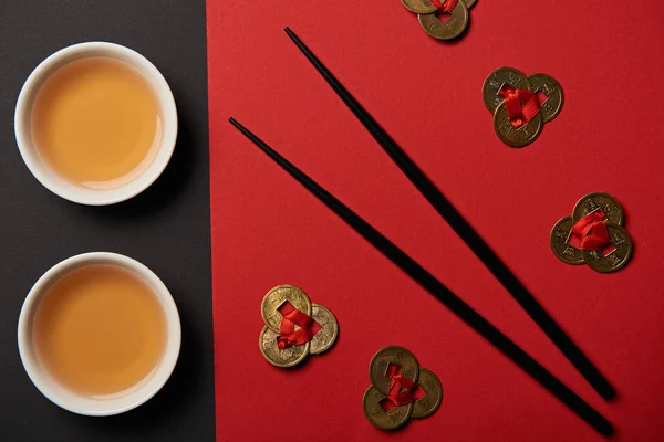 Vue du dessus des tasses à thé, des pièces de feng shui et des baguettes sur fond rouge et noir — Photo de stock