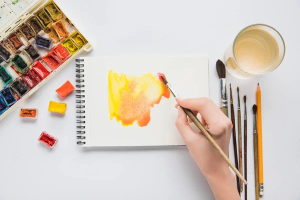 Vue du dessus des mains féminines dessin dans l'album avec aquarelle peintures et pinceau — Photo de stock