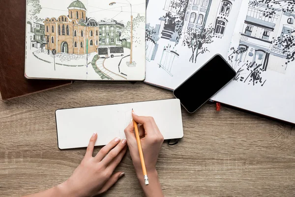 Vista dall'alto delle mani delle donne che disegnano su carta, album con vernici e smartphone su sfondo di legno — Foto stock