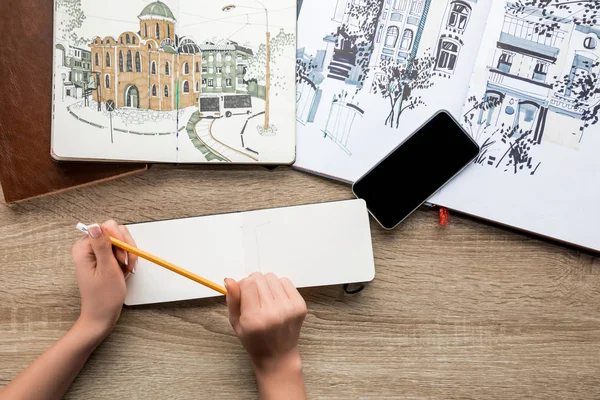 Вид сверху на женские руки с карандашом, альбомы с красками и смартфоном на деревянном фоне — стоковое фото