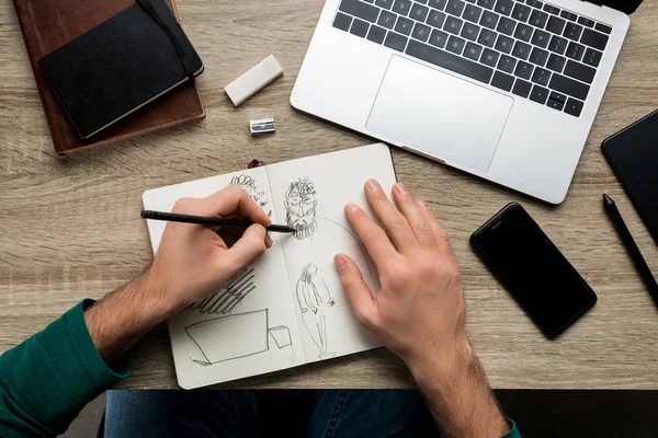Вид сверху рук людей, рисующих на альбоме и смартфоне рядом с ноутбуком на деревянном столе — стоковое фото