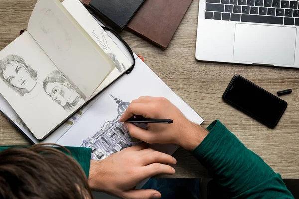 Overhead back view of man drawing in album on wooden table next to laptop and smartphone — Stock Photo