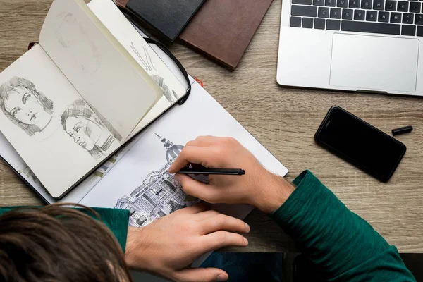 Vue aérienne arrière du dessin de l'homme dans l'album sur une table en bois à côté de l'ordinateur portable et du smartphone — Photo de stock