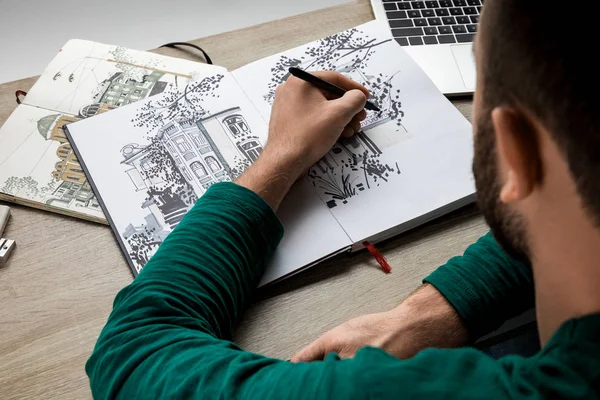 Vue arrière du dessin de l'homme dans l'album sur la table en bois à côté de l'ordinateur portable — Photo de stock