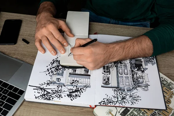 Vista dall'alto di mani mans disegno in notebook su tavolo di legno accanto a album e gadget — Foto stock