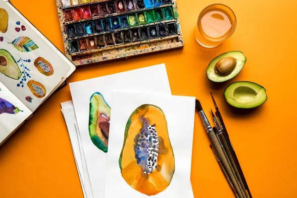 Top view of colored paints, paintbrushes, sketchbook, drawings with avocado and papaya on yellow background — Stock Photo